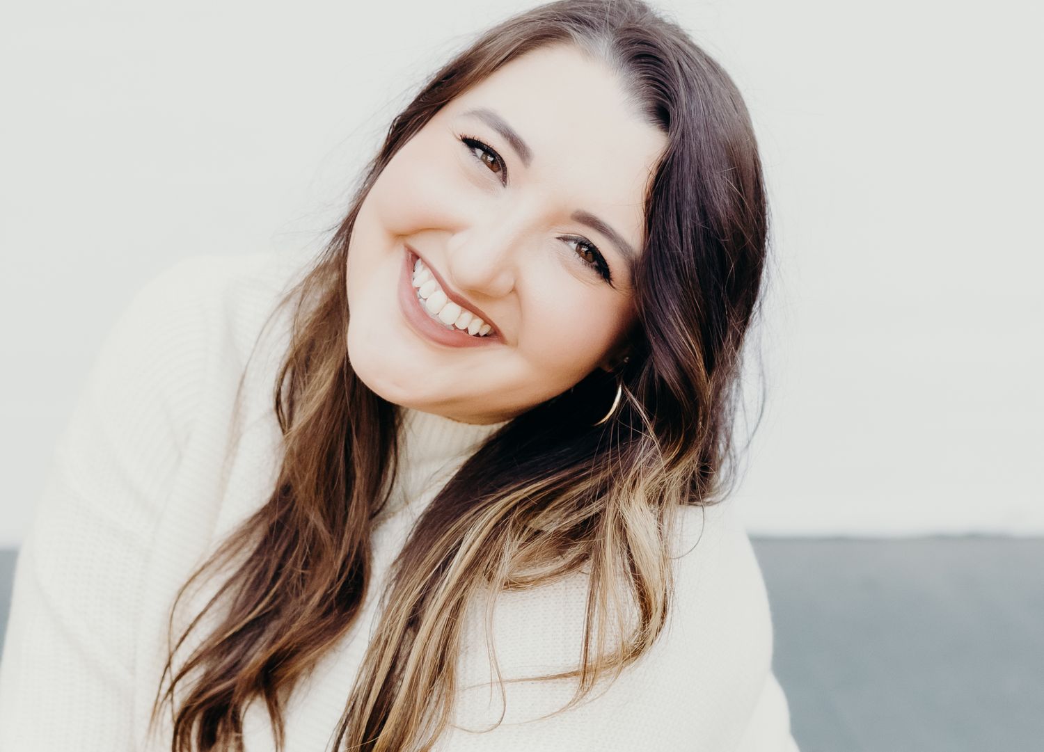 A person with long brown hair and a bright smile wearing a white sweater poses against a light background.