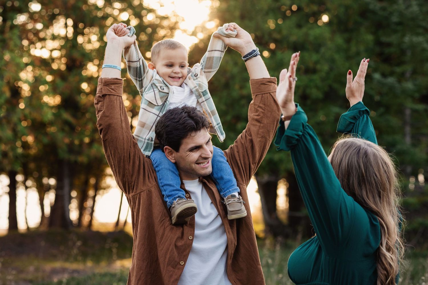A family celebrates together outdoors at sunset with their arms raised joyfully in a wooded setting.