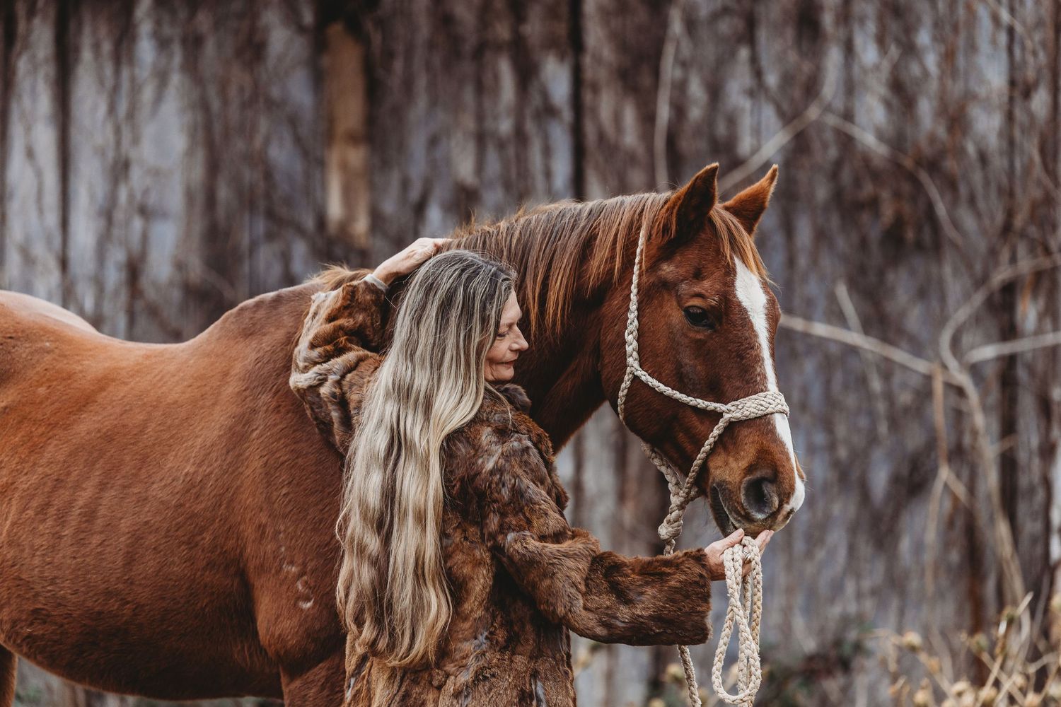 Blonde woman in fur coat standing close with chestnut horse wearing white halter outdoors.