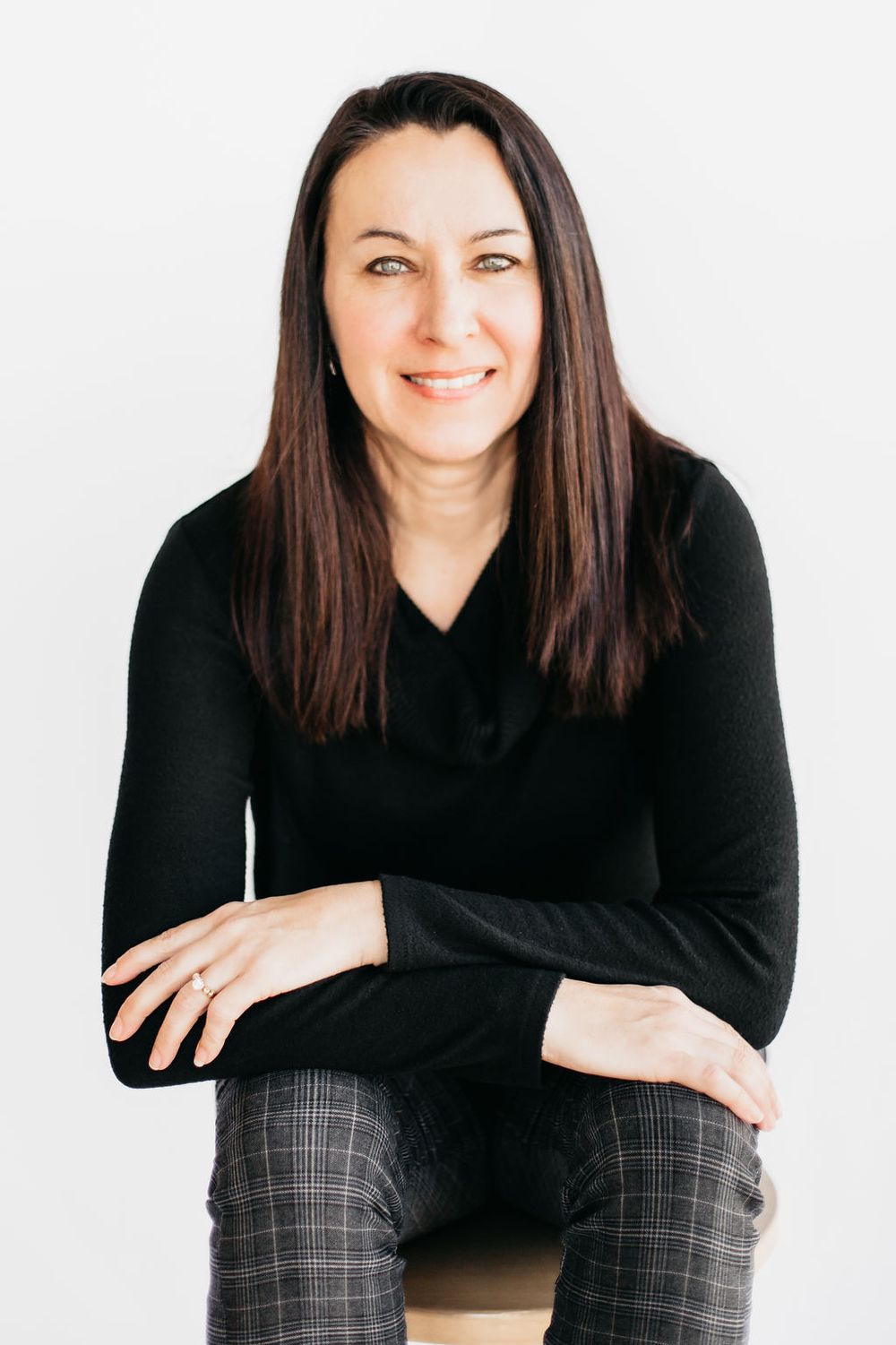 A smiling woman with dark hair wearing a black top and plaid pants sits with arms crossed against a white background.
