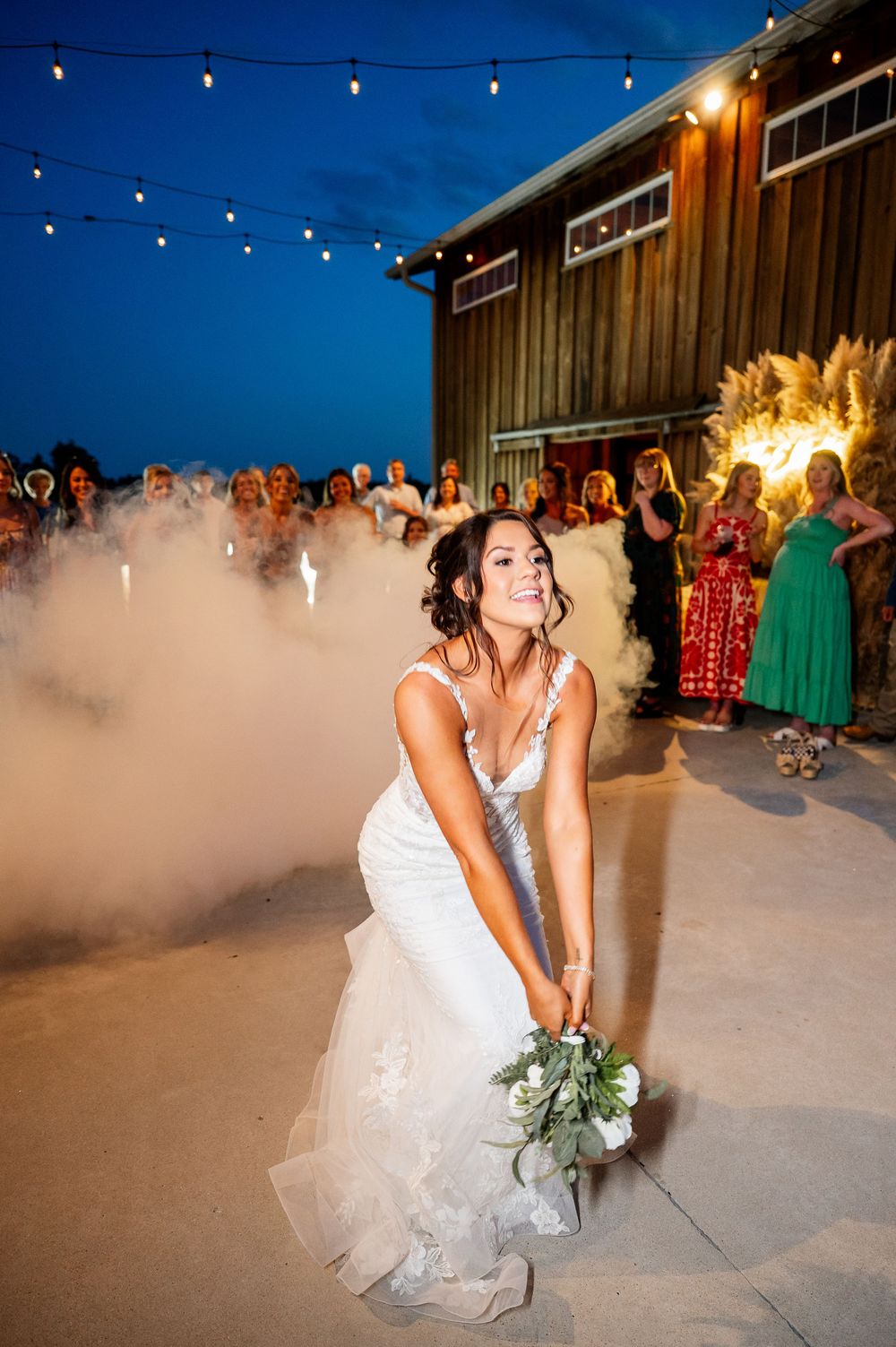 A bride dances in a cloud of smoke under string lights during an evening wedding reception.
