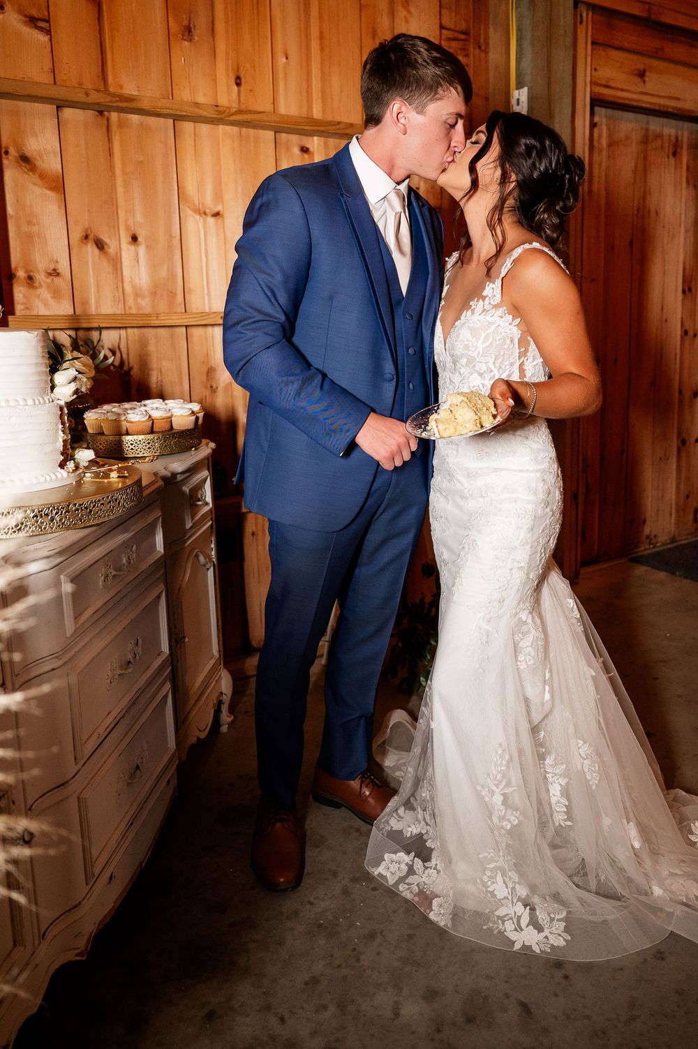 Wedding couple shares a fun cake-feeding moment in a rustic barn venue with wooden panel walls.
