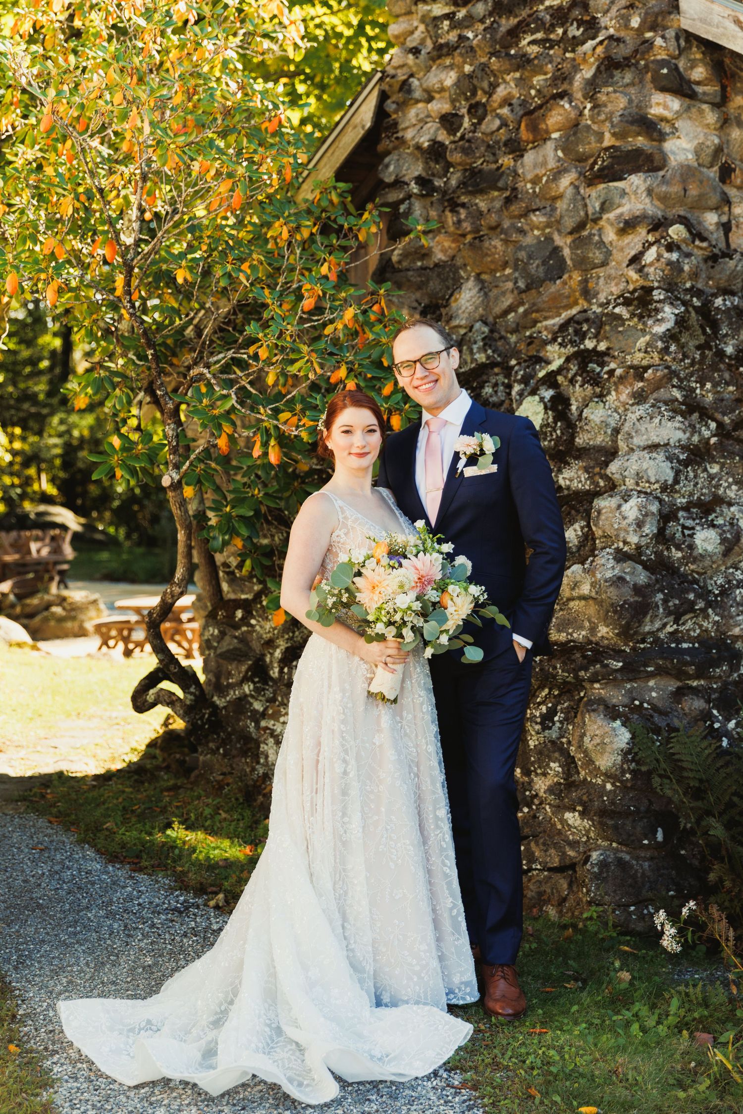 A couple poses against a rustic stone wall with orange blossoms in the background.