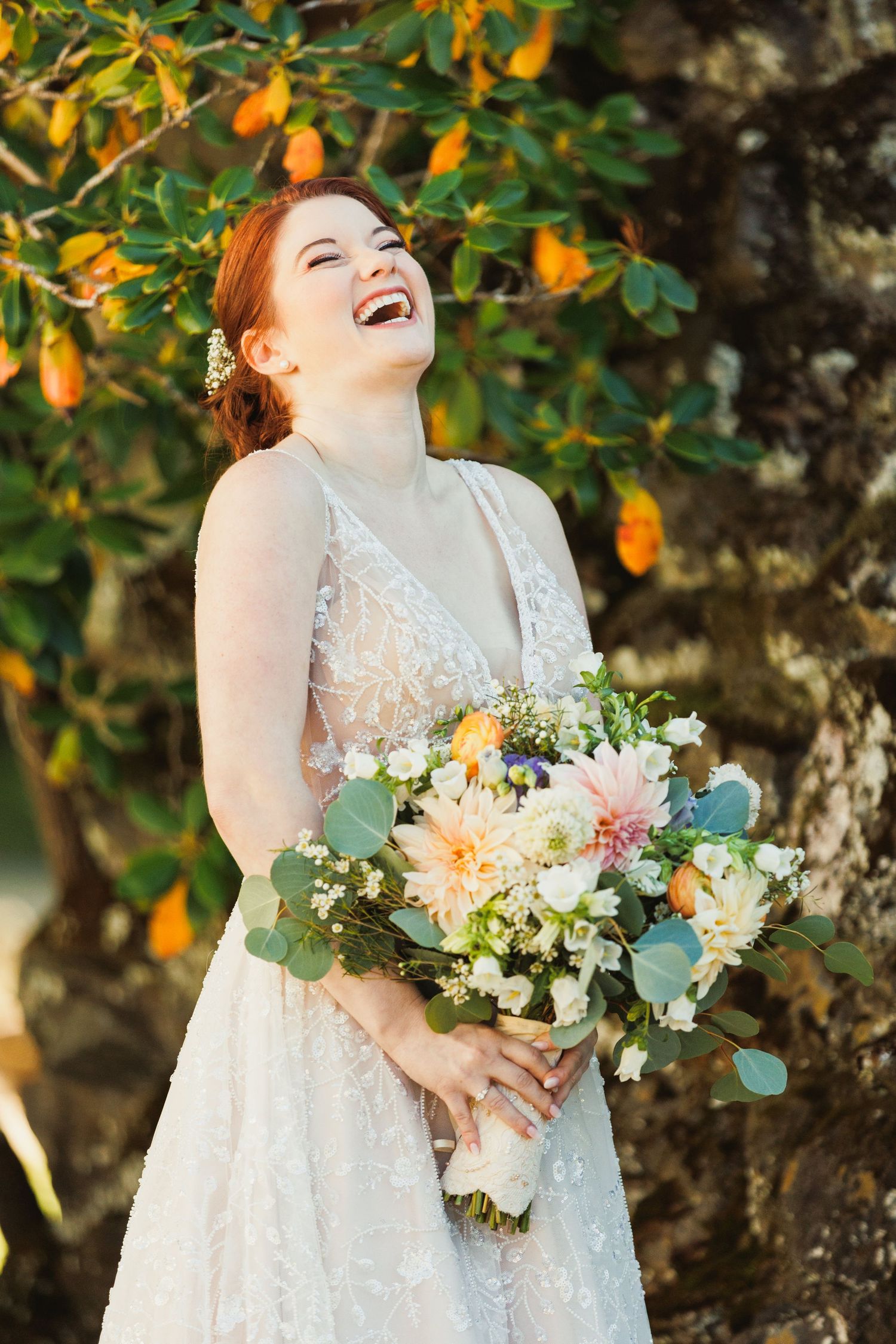 A joyful bride laughs while holding a pastel bouquet under orange blossoms.