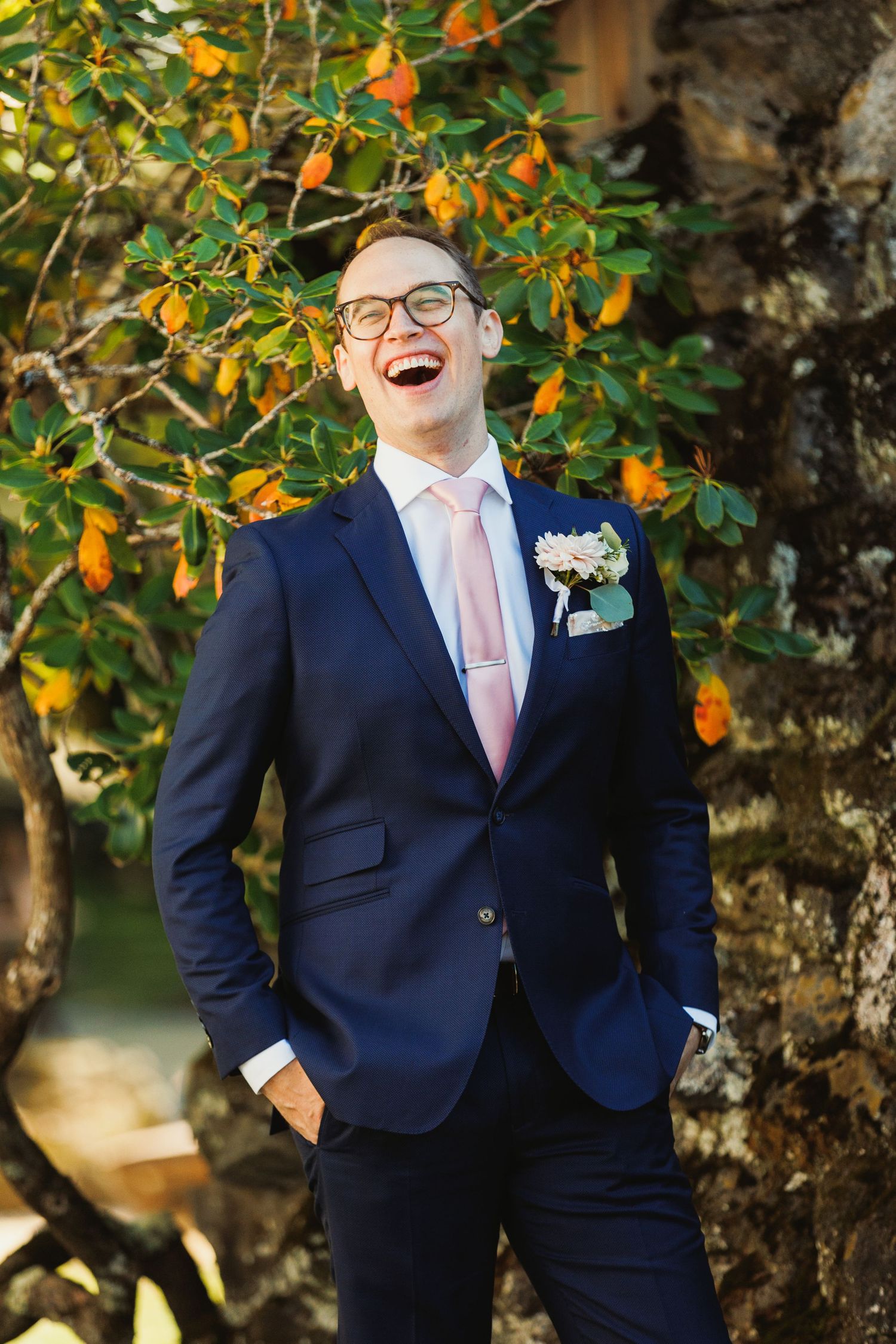 A groom laughs joyfully in front of orange blossoms wearing a navy suit and pink tie.