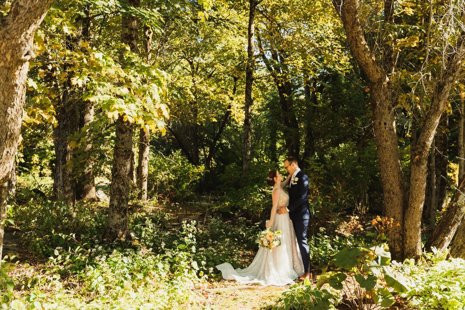 A couple stands together in a sunlit forest wearing elegant wedding attire.