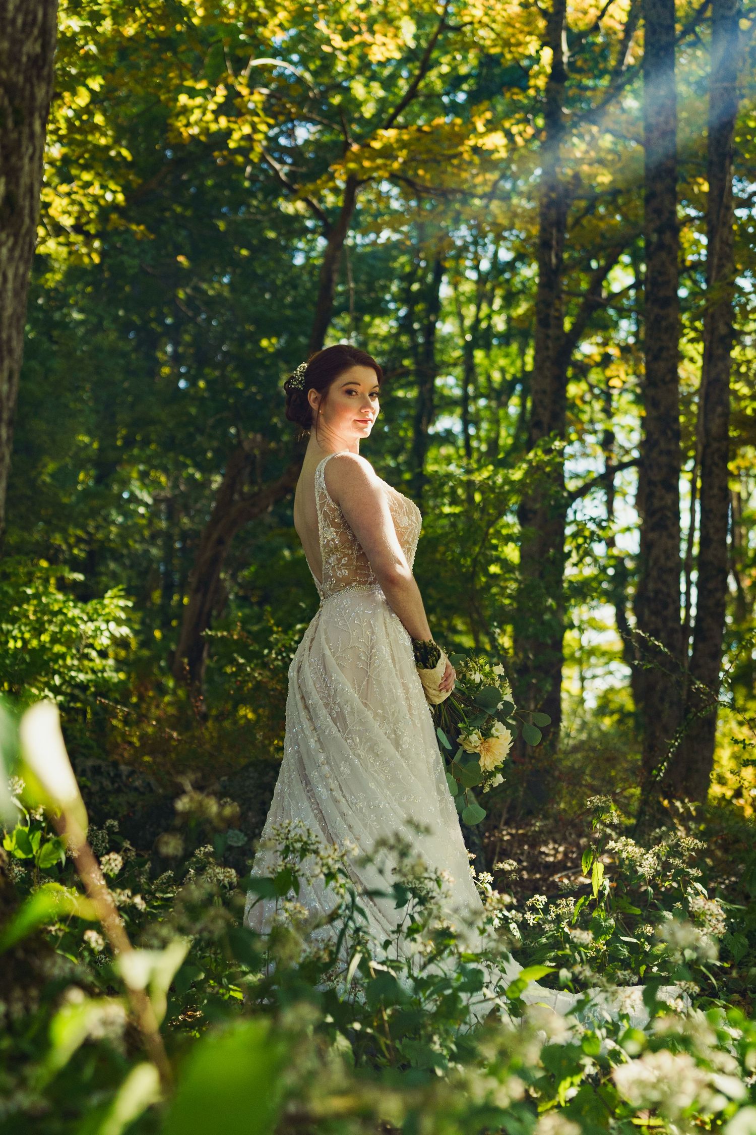 A bride in a lace wedding gown stands in a wooded area with dappled sunlight.