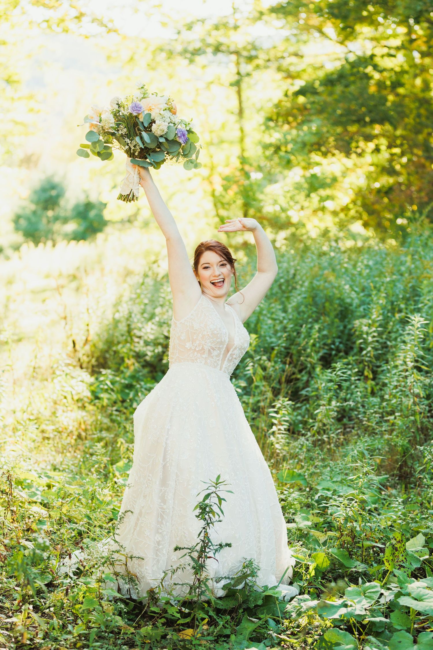 Joyful bride holding bouquet aloft in sunlit forest setting.