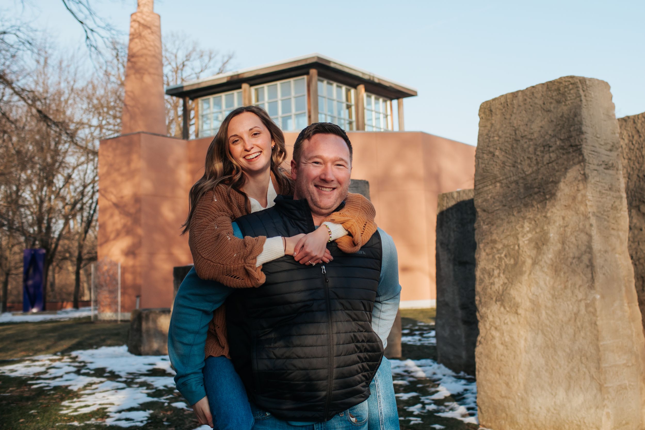 A couple embraces outdoors during sunset near stone monuments on a winter day.