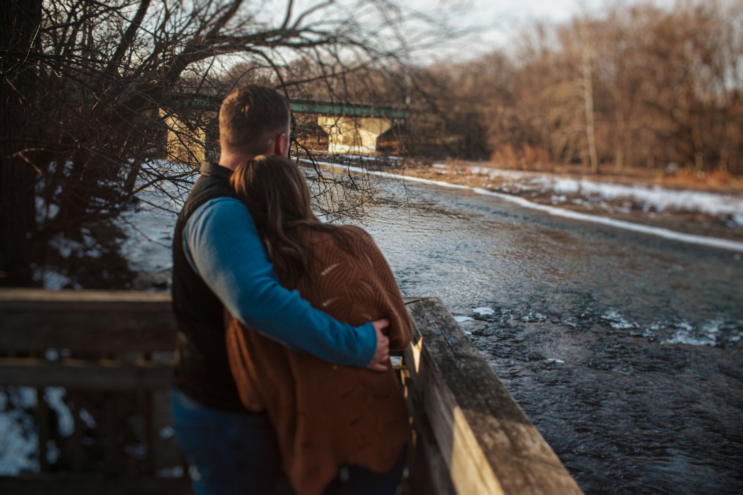 A couple shares an intimate moment on a snowy bridge at dusk.