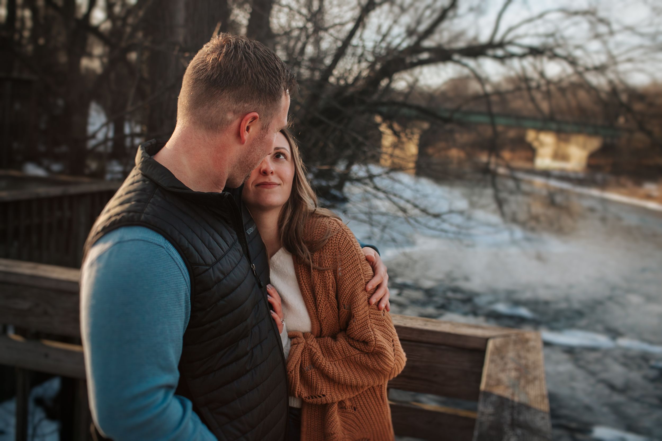 A couple shares an intimate moment on a bridge overlooking a river during sunset.