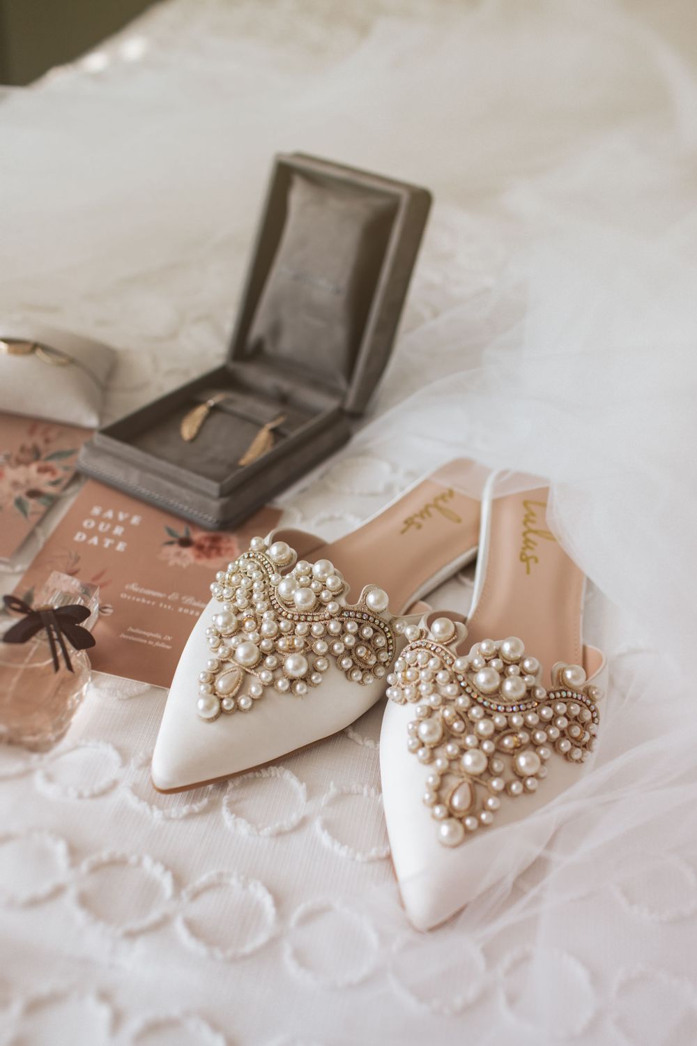 Elegant bridal shoes adorned with pearl embellishments photographed on white lace fabric with a gray jewelry box.