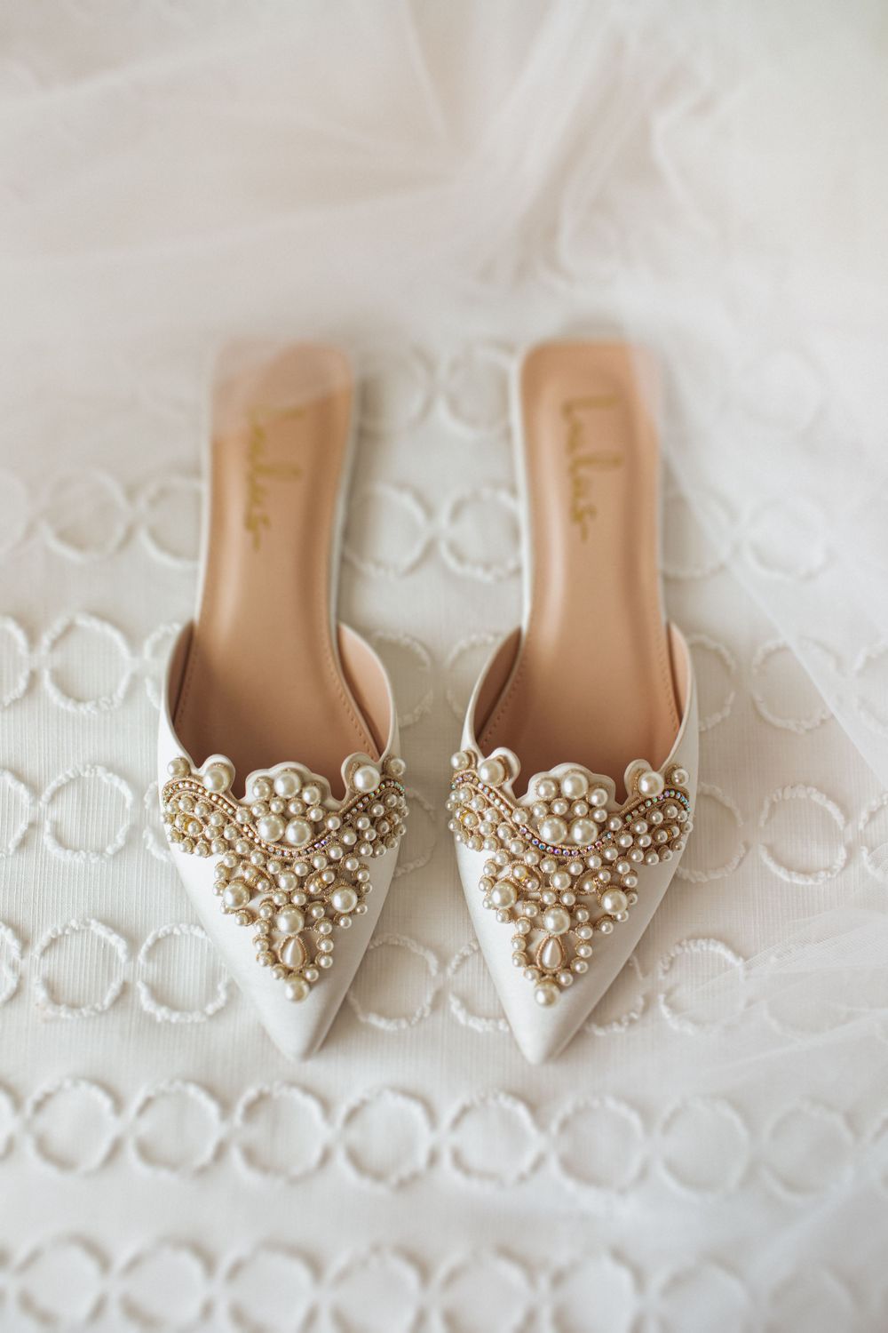 Elegant pearl-embellished bridal shoes with pointed toes resting on white embroidered fabric.