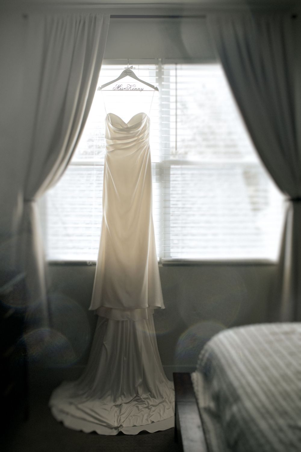 An elegant white wedding dress hangs from a hanger in front of a window with flowing curtains.
