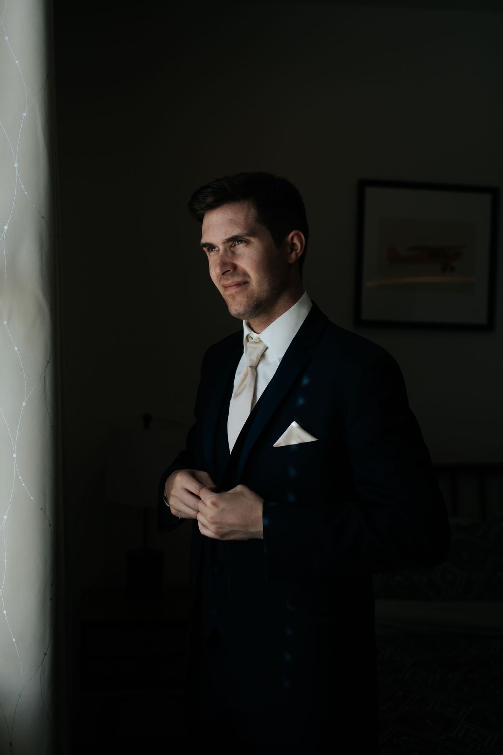 A formal black suit and tie ensemble is worn in dramatic window lighting.