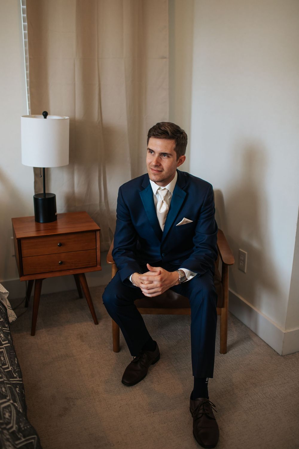 A person in a navy suit sits thoughtfully on the floor against a beige wall with wooden side tables.