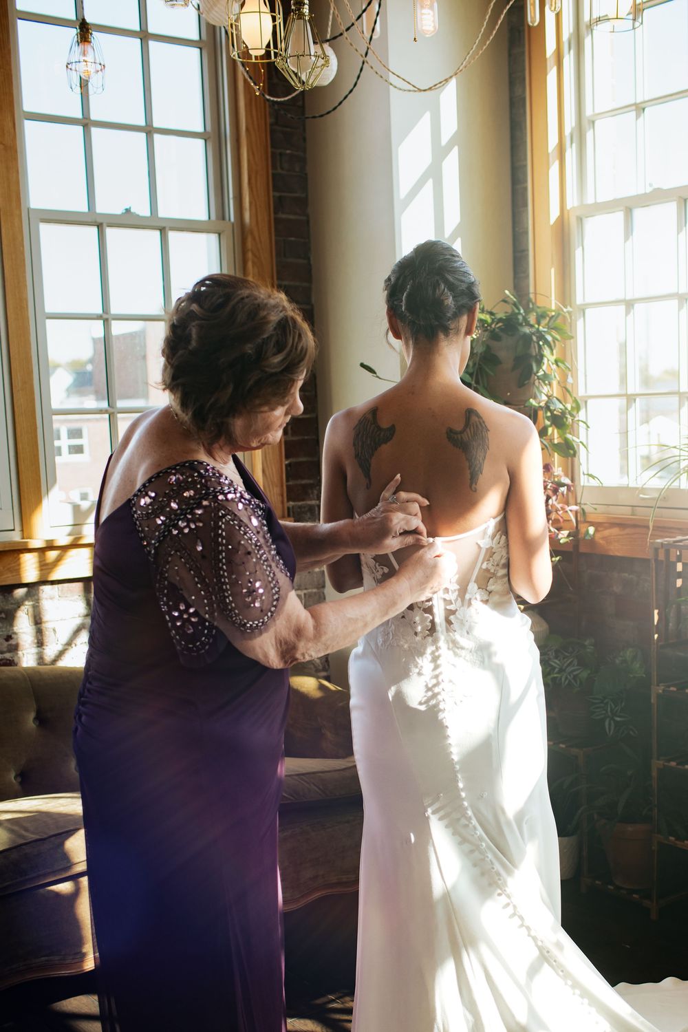 A wedding dress fitting in a sunlit room with a purple-clad helper making final adjustments to the white gown.