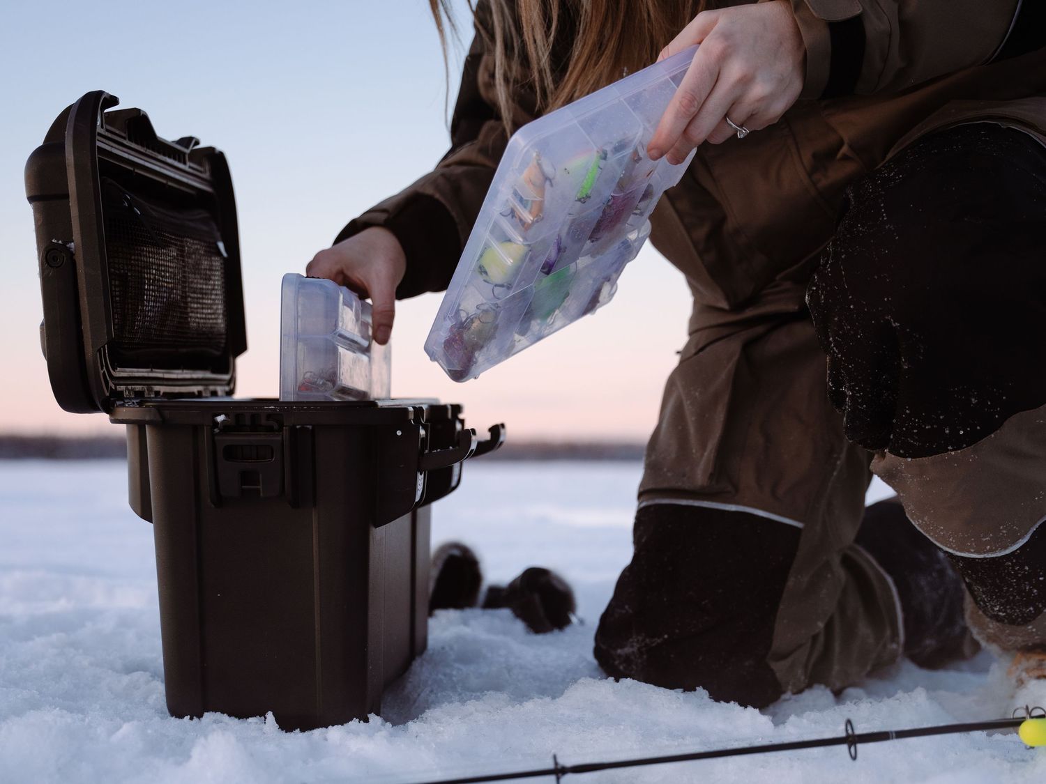 Hands holding ice fishing equipment against snowy winter landscape at sunset.