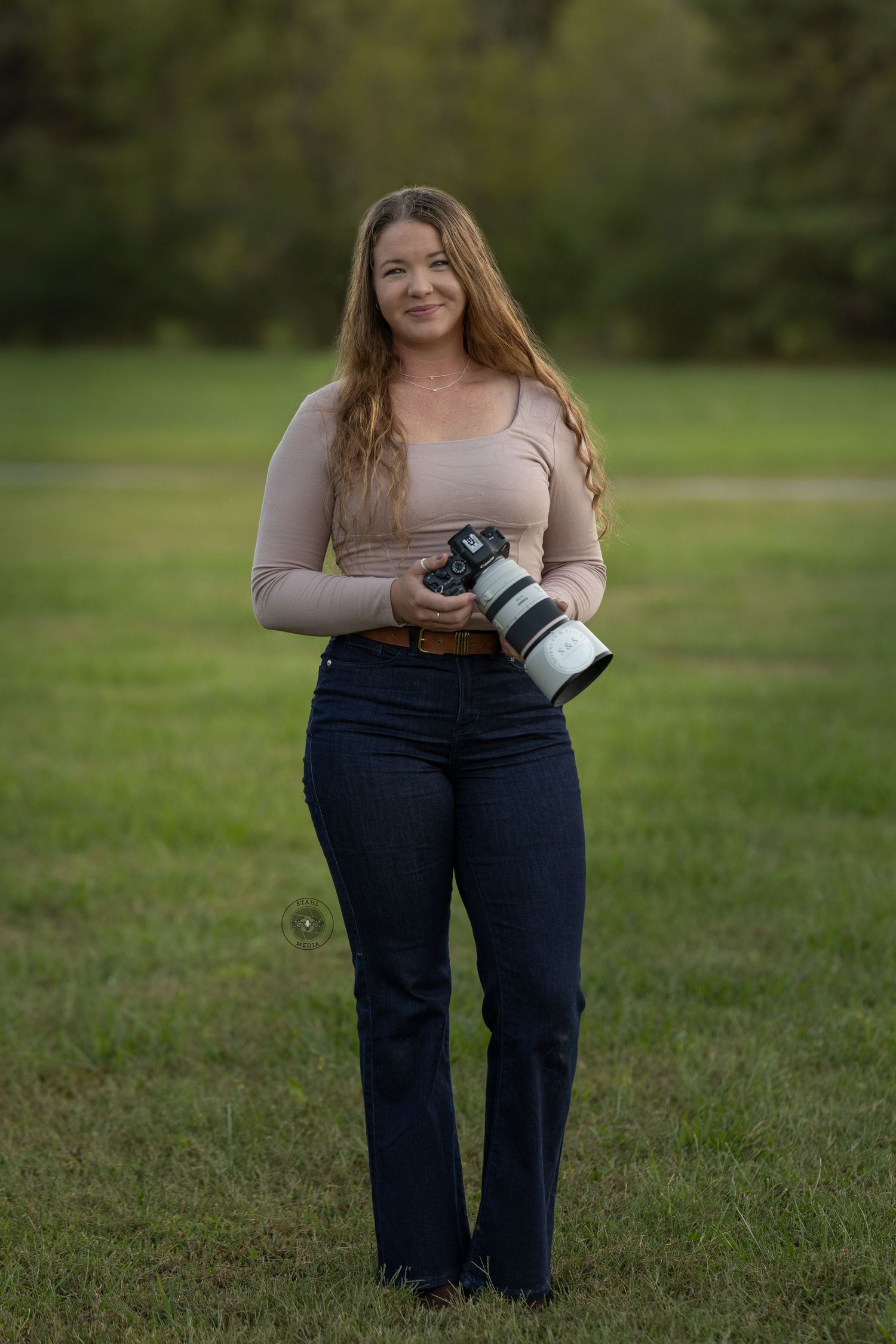 A photographer stands in a grassy field holding a professional camera while wearing jeans and a beige top.