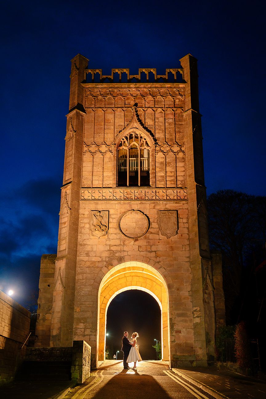 Dramatic nighttime wedding photos at historic stone archway illuminated in warm lighting.