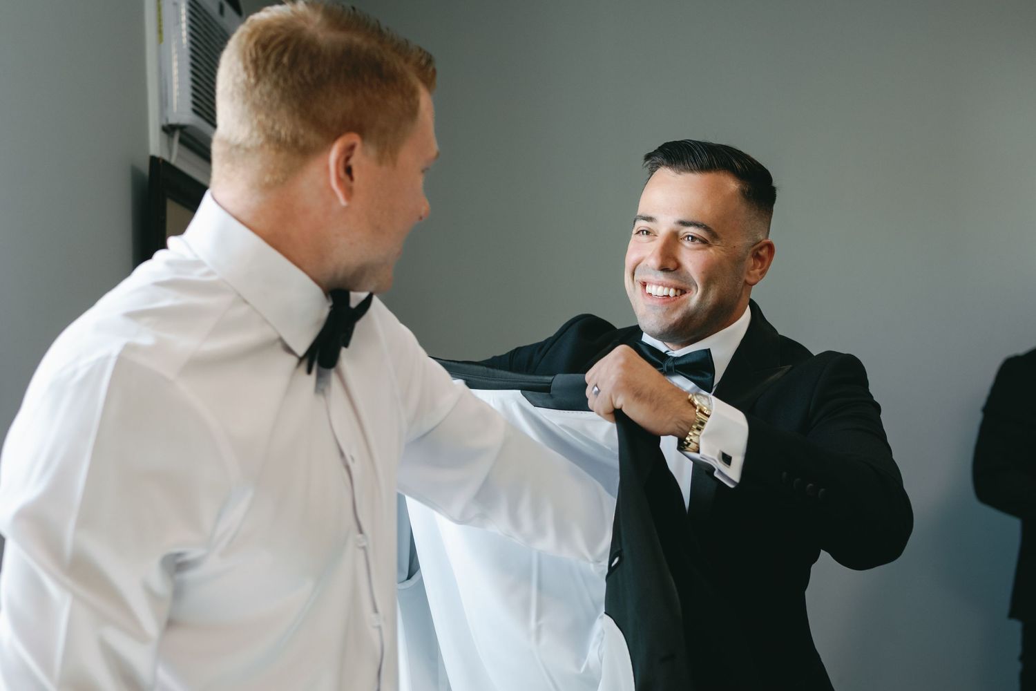 A romantic couple in dark suits share a tender moment on a balcony during a wedding celebration.