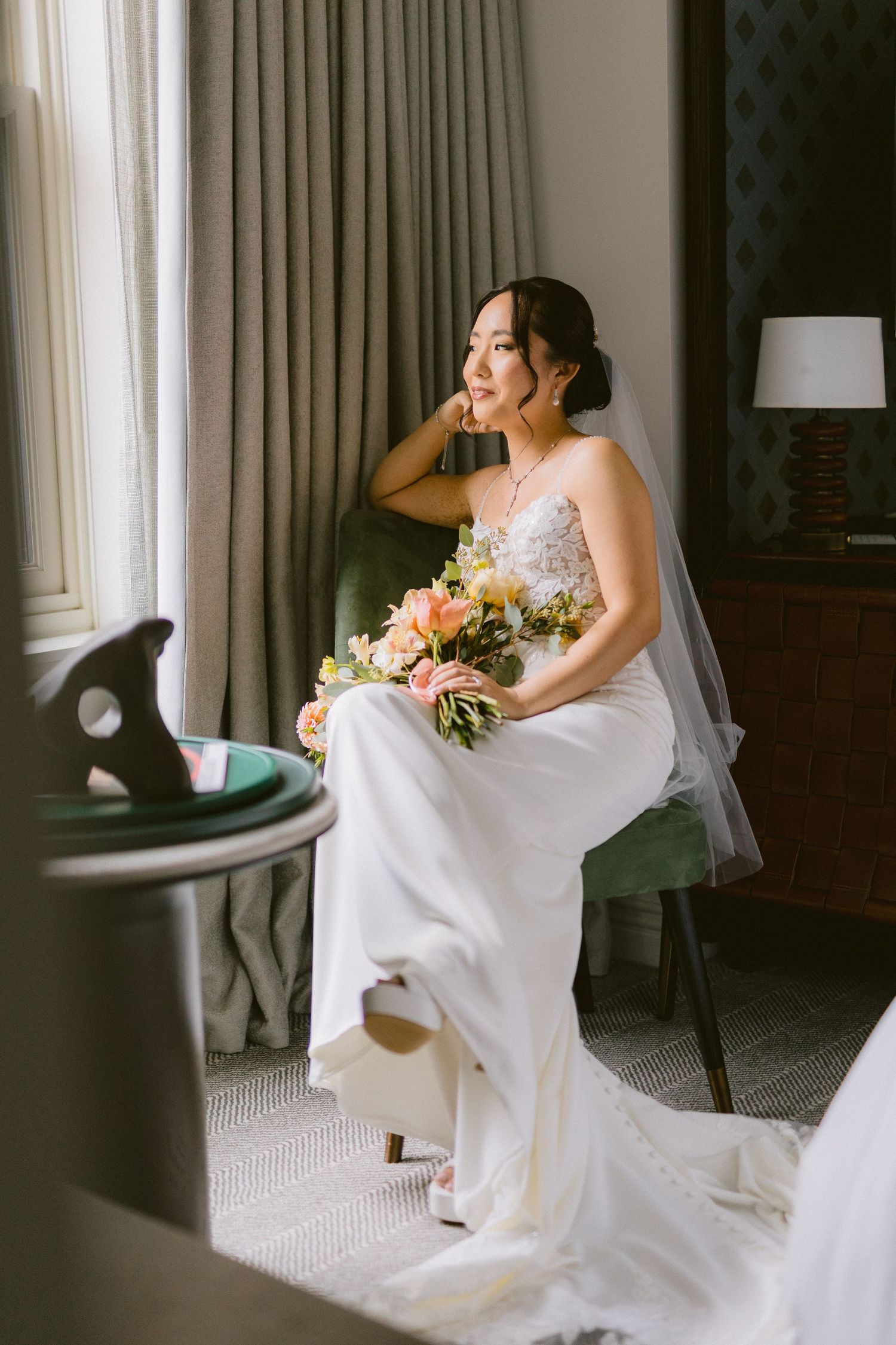 A bride in an elegant white gown sits thoughtfully by a window holding a pink and white floral bouquet.