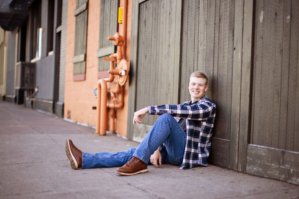 Person in blue jeans and plaid shirt leans against concrete wall in urban setting.