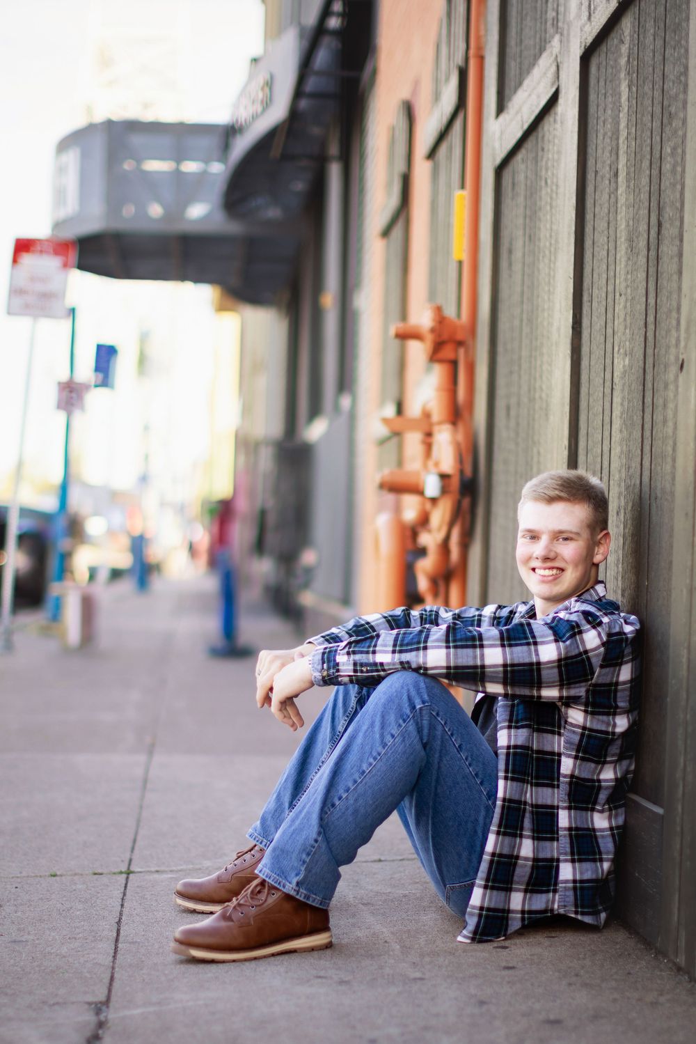 Casual portrait of someone sitting on sidewalk wearing plaid shirt and jeans in urban setting.