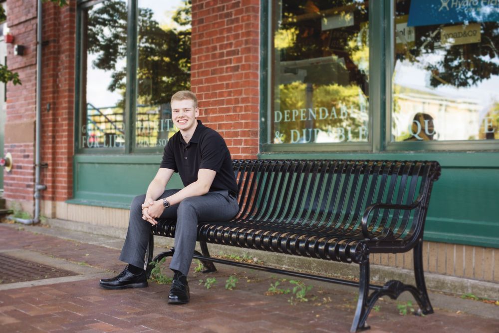 A person sits on a black metal bench outside a brick building with green trim and windows.