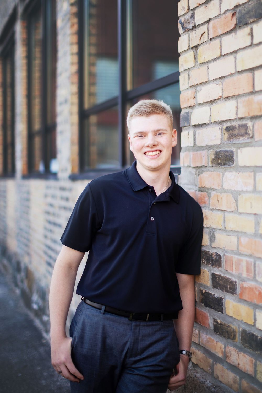 Professional portrait of someone in a navy polo shirt and dress pants standing against a brick wall background.