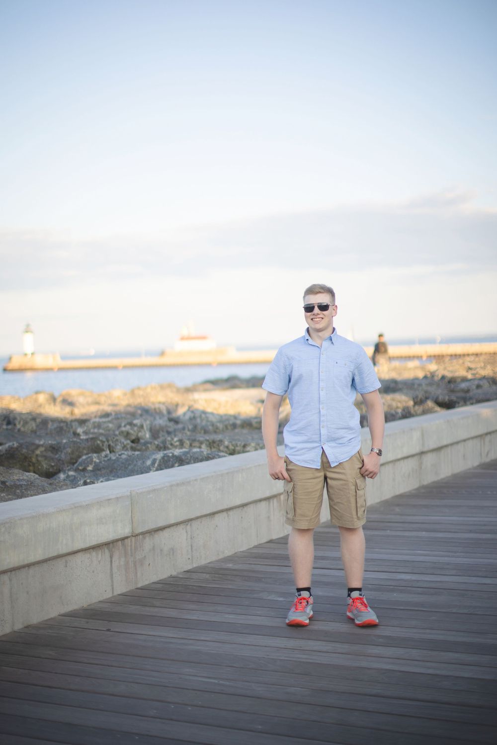 A person in casual attire stands on a wooden boardwalk during sunset with a scenic coastal backdrop.