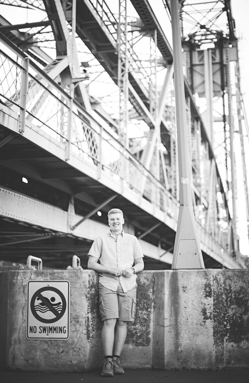 Black and white photo series showing a person walking along a concrete wall beneath a bridge structure with a No Swimming sign.