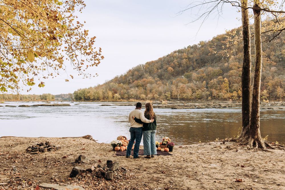 Couple embraces facing autumn river with colorful hillside and pumpkin decorations.