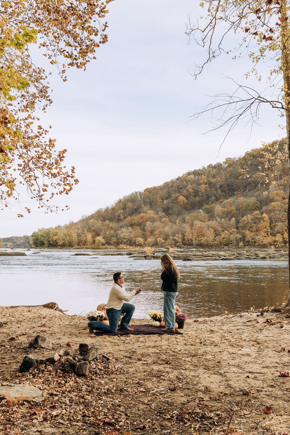 Man proposes to woman by riverside with autumn flowers and pumpkin decorations.