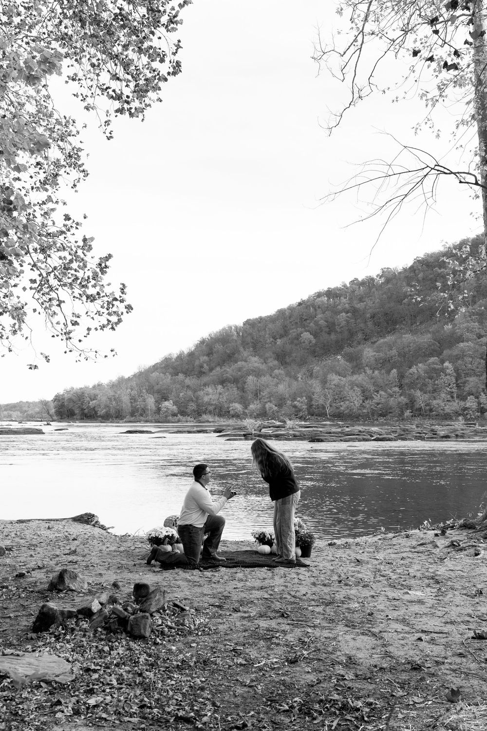 Black and white photo of riverside marriage proposal with wooded hill backdrop.