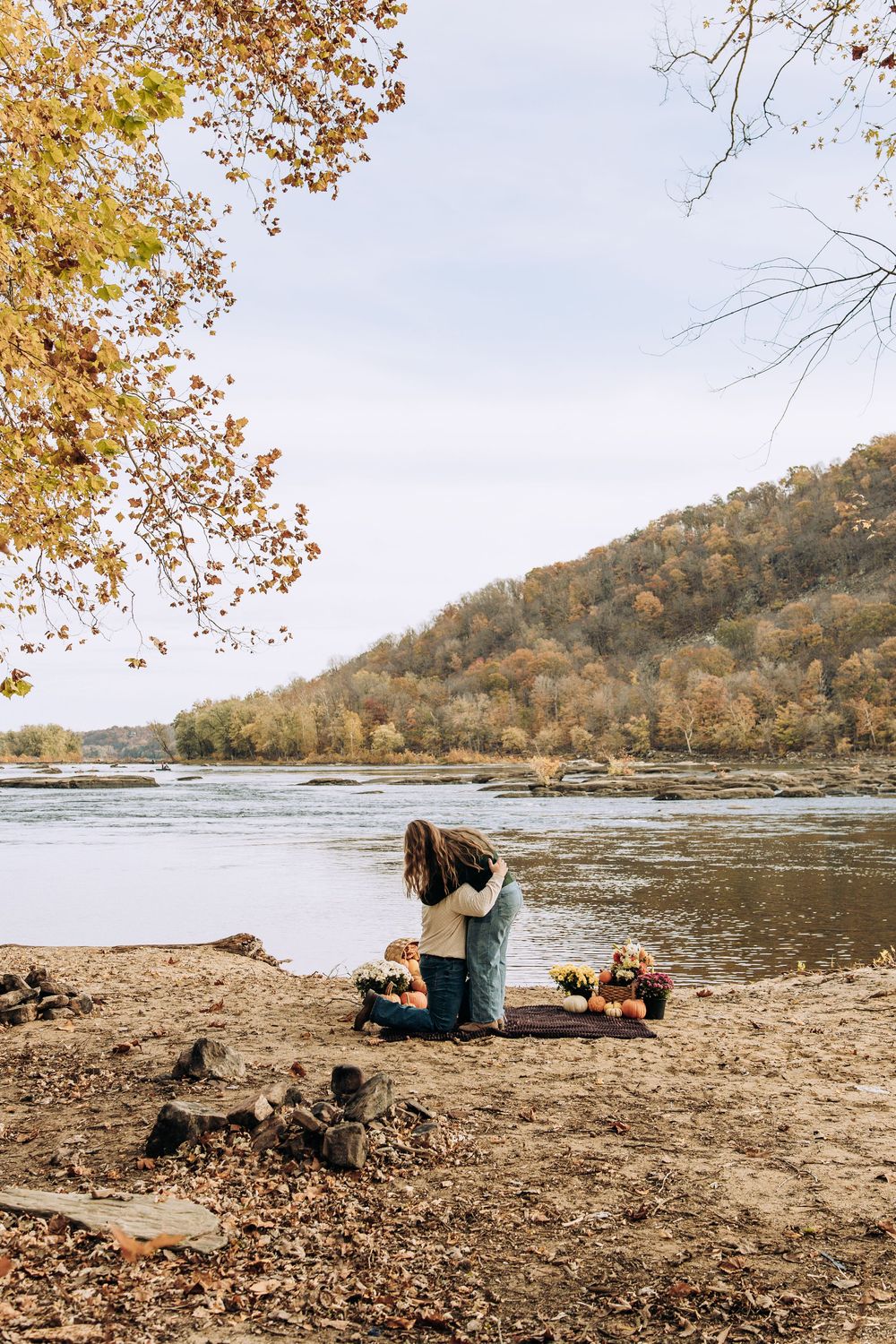 Couple embraces on knees by river after proposal surrounded by fall flowers.