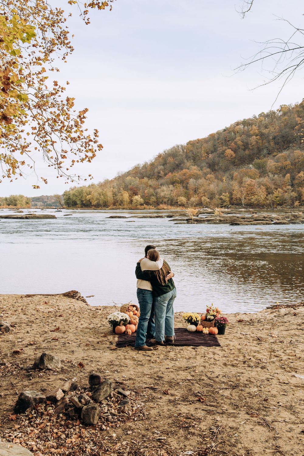 Couple hugs facing autumn river with pumpkins and flowers on blanket behind them.