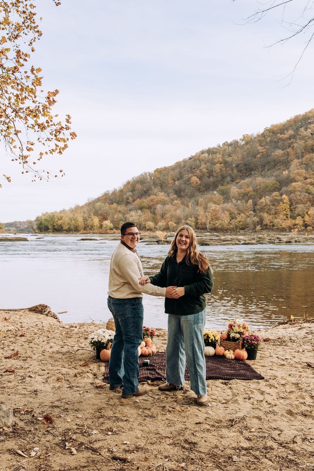 Smiling newly engaged couple holds hands by autumn river with pumpkin display.