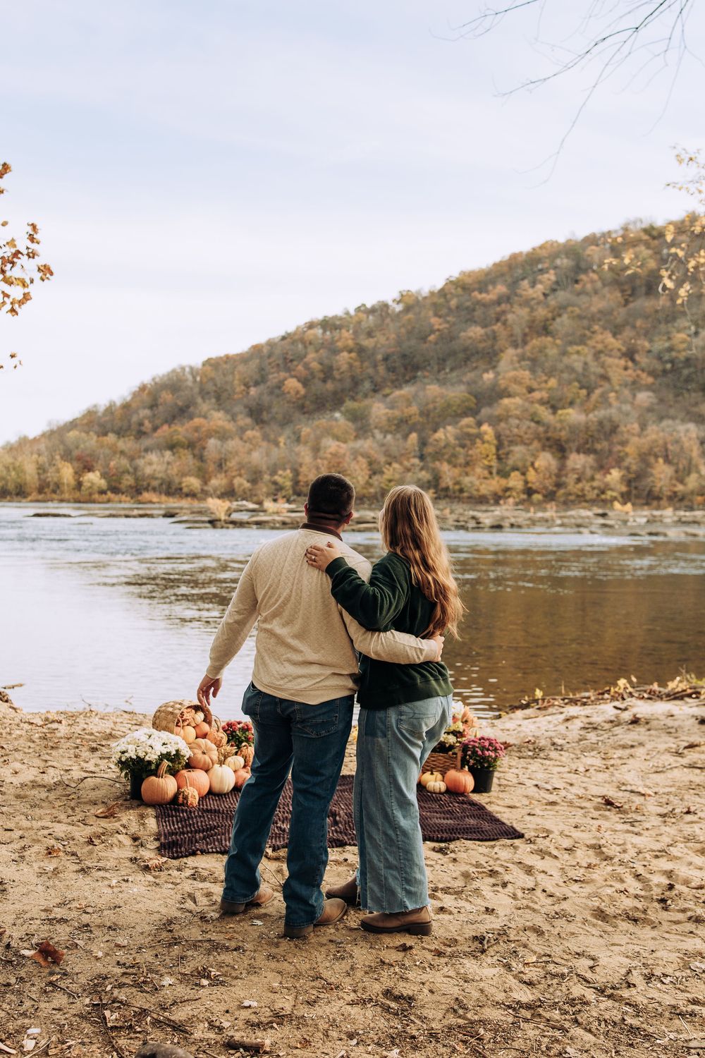 Couple embraces by scenic autumn river with pumpkins and flowers on blanket.