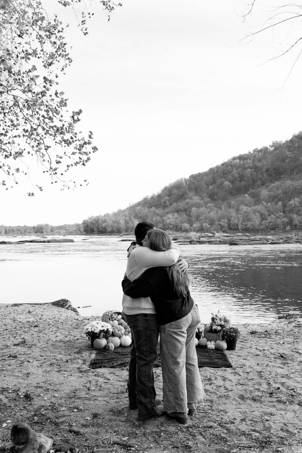 Black and white photo of couple hugging by river after proposal with pumpkins.