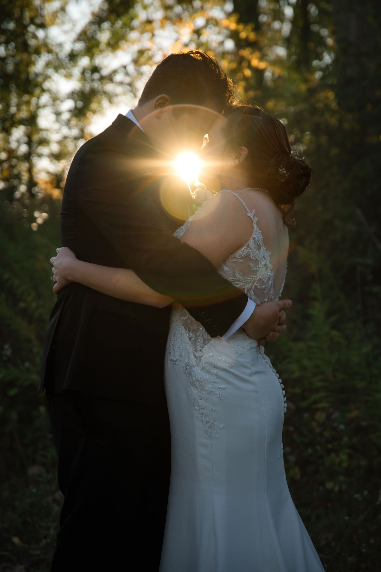 A couple in wedding attire embraces in a wooded setting with sunlight streaming through the trees creating a romantic silhouette.