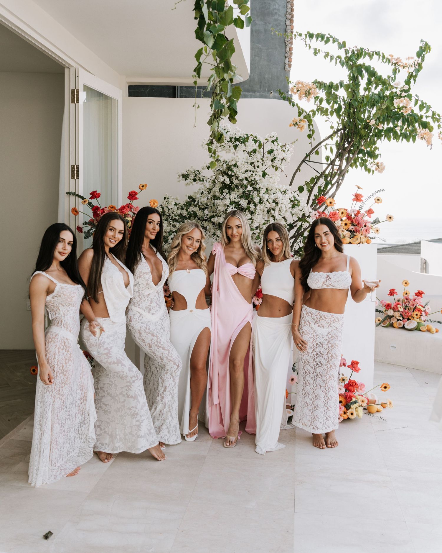 A group in white dresses and one in pink pose together in a bright corridor with flowering plants.