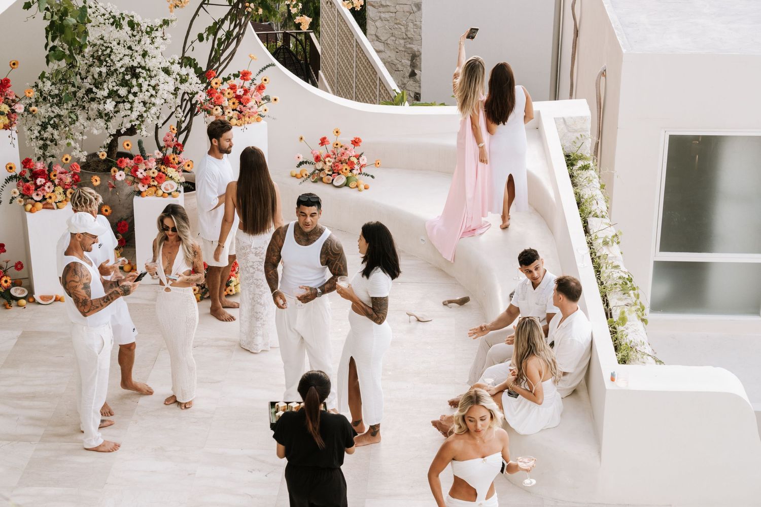 A group of people in white outfits gather for a celebration on a bright Mediterranean-style patio with coral floral decor.