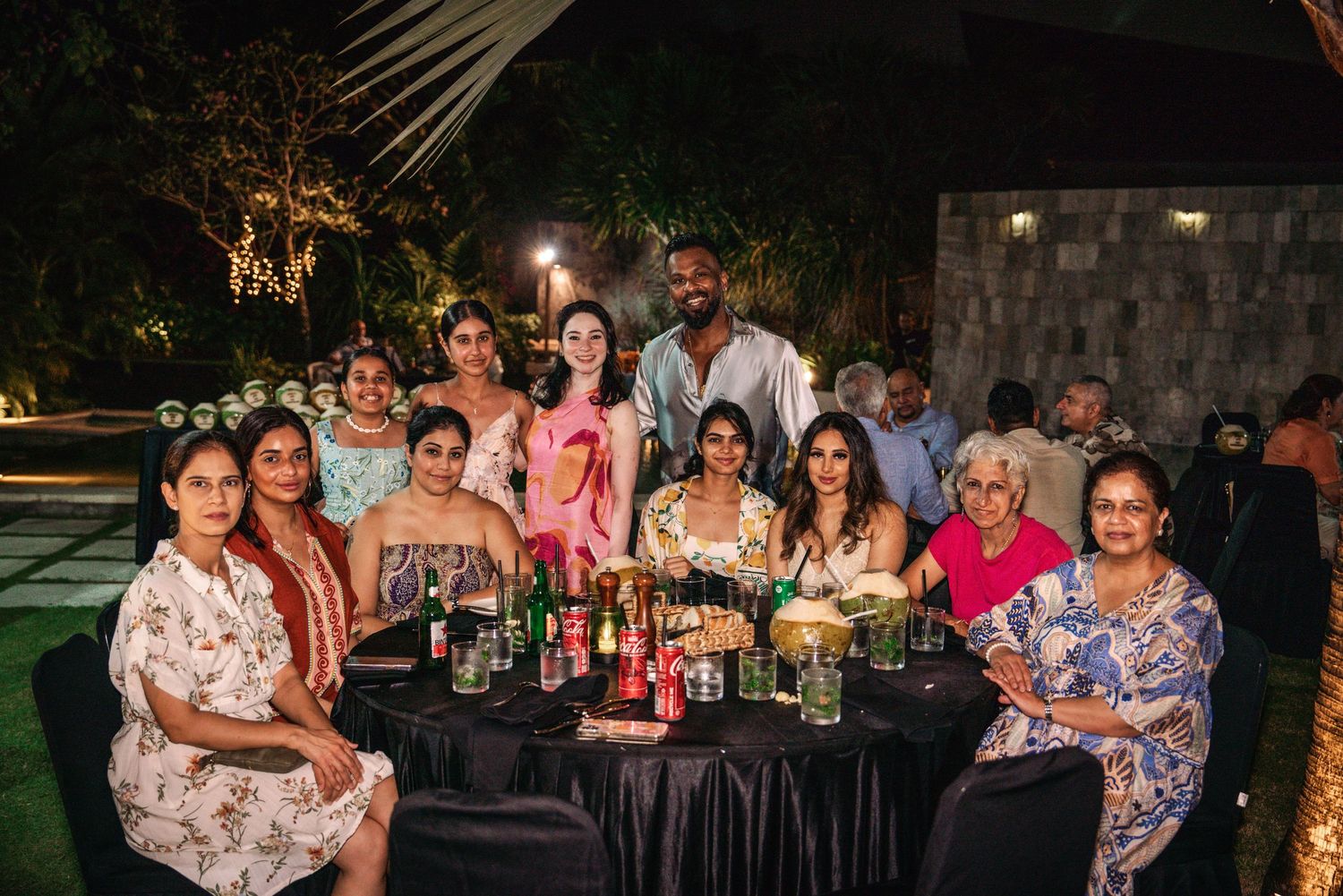 A group of people sit at a long dining table outdoors at night during a festive gathering.