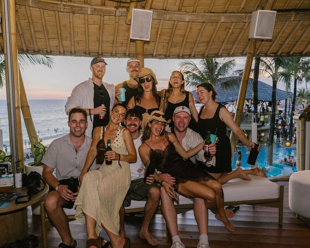 Group of friends posing together at a tropical beach bar with ocean views, palm trees, and a pool at sunset.