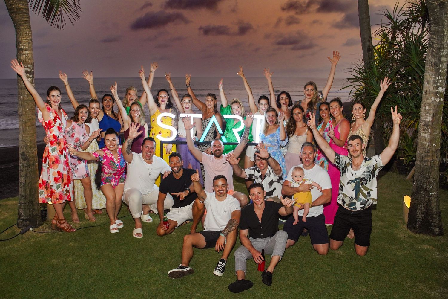 Group of people jumping and posing together outdoors at sunset with palm trees in the background.
