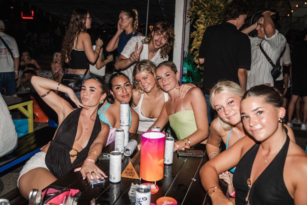 A group of party-goers enjoying drinks and socializing at a nightclub with festive lighting in the background.