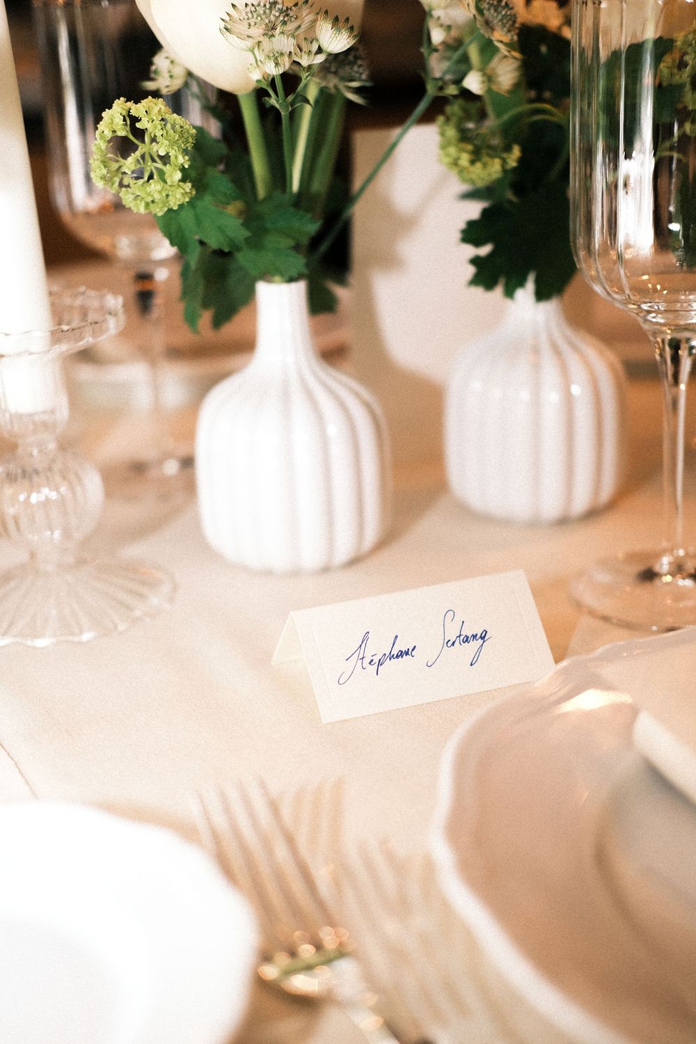 Elegant table setting with white ribbed vases holding flowers and a handwritten place card on cream tablecloth.