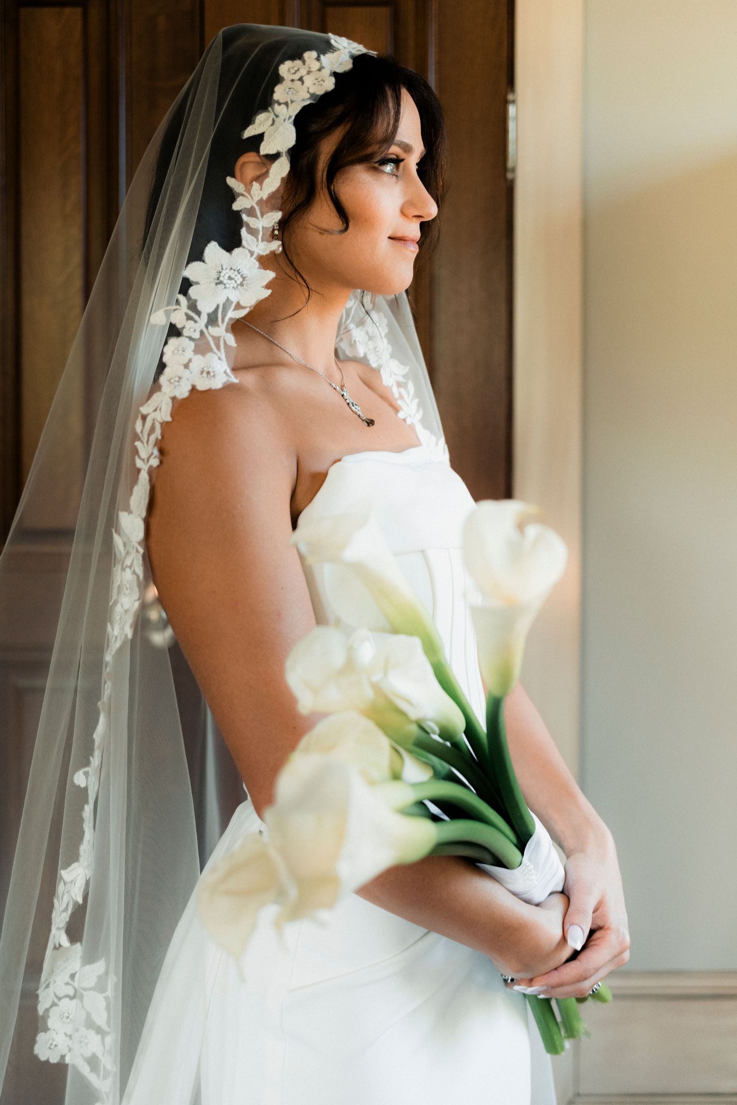 Bride in strapless white gown and lace veil holding white calla lily bouquet by door.