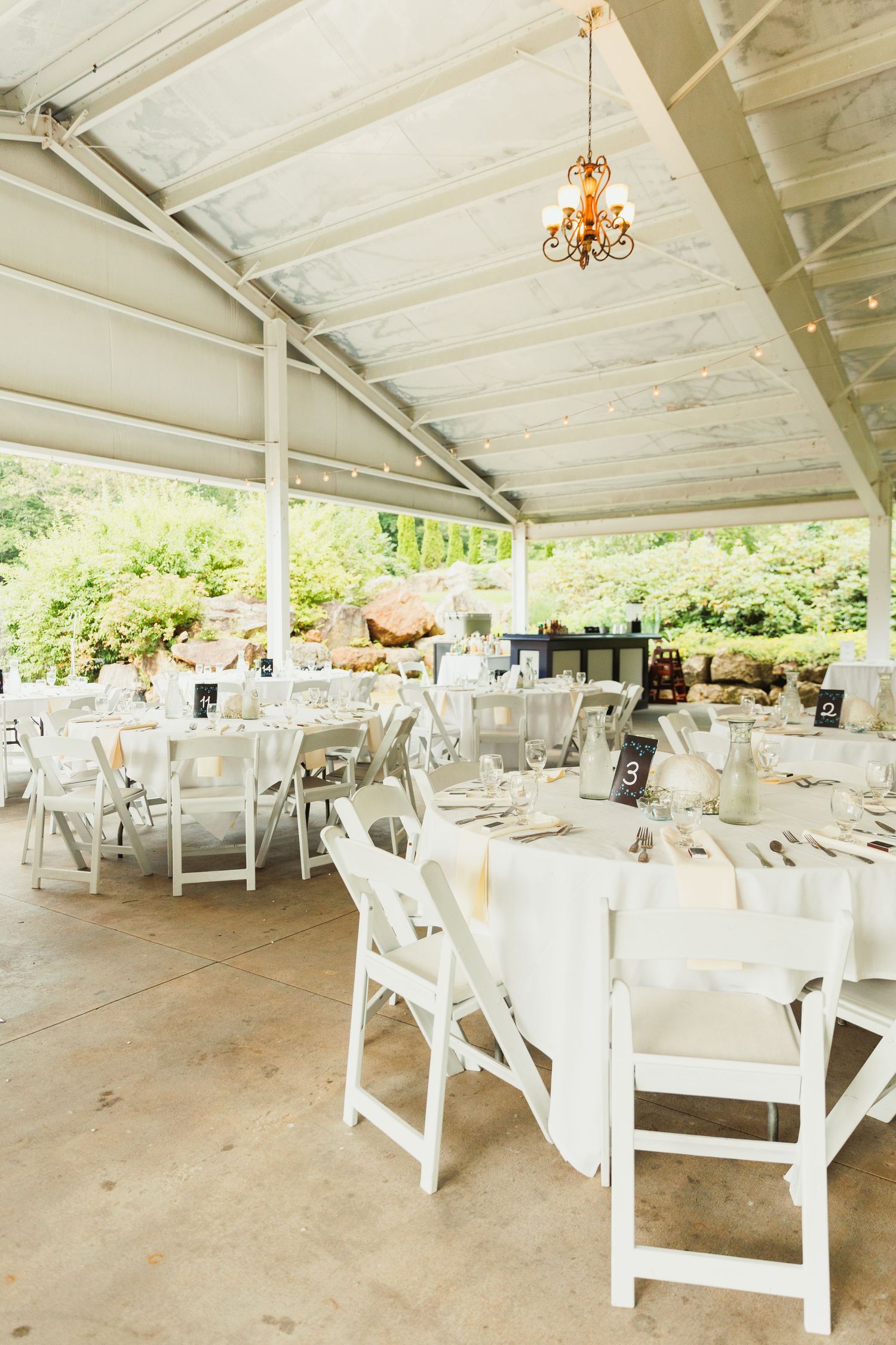 An elegant outdoor wedding reception venue with white chairs and tables under a covered pavilion.